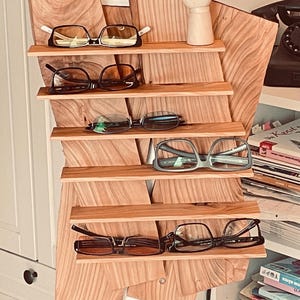 Könnte beinhalten: Ein Brillenständer aus Holz mit sechs Regalen, auf denen jeweils eine Brille liegt. Die Brillen variieren im Stil, darunter schwarze, silberne und transparente Rahmen. Der Ständer ist aus hellem Holz gefertigt und an der Wand befestigt.
