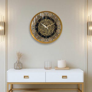 May include: A gold framed clock with black and gold Arabic numerals and a decorative design. The clock is hanging on a white wall with gold trim. A white table with gold legs is in the foreground.