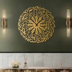 May include: A gold-colored, circular wall clock featuring Arabic calligraphy. The clock has black hands and Roman numerals at the top. The clock is mounted on a dark green wall, with two gold sconces on either side. Below the clock is a marble-topped console.