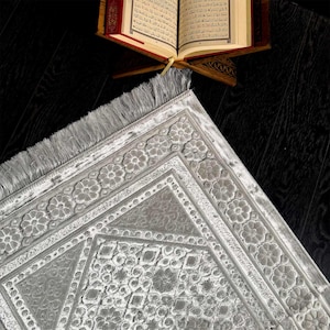May include: A gray prayer rug with a floral border and geometric center design. The rug has a fringed edge and is placed on a dark wooden surface. An open book rests on a wooden stand in the background.