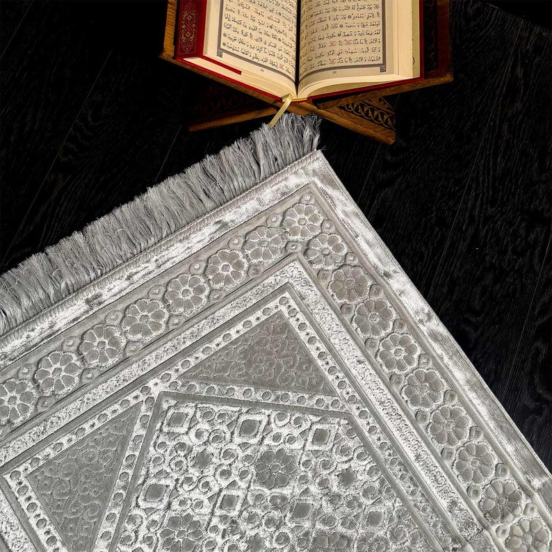 May include: A gray prayer rug with a floral border and geometric center design. The rug has a fringed edge and is placed on a dark wooden surface. An open book rests on a wooden stand in the background.