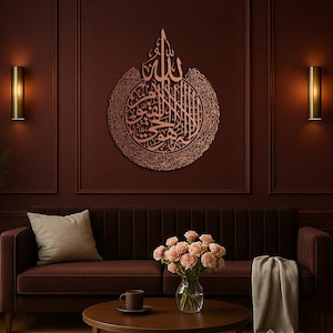 May include: A copper-colored, circular wall art piece featuring Arabic calligraphy. The artwork is mounted on a dark red wall above a brown velvet sofa. The room is decorated with sconces and a coffee table with a vase of pink roses.
