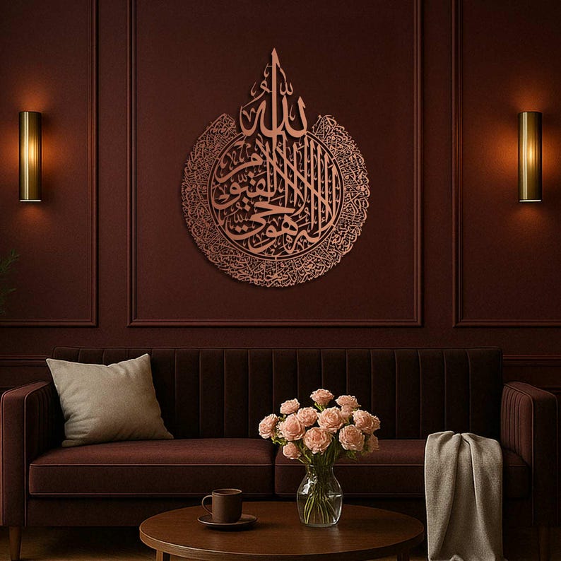 May include: A copper-colored, circular wall art piece featuring Arabic calligraphy. The artwork is mounted on a dark red wall above a brown velvet sofa. The room is decorated with sconces and a coffee table with a vase of pink roses.