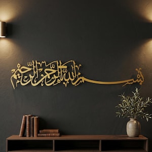May include: A gold-colored metal wall art piece featuring Arabic calligraphy of the Basmala. The script is mounted on a dark gray wall above a wooden shelf holding a stack of books and a ceramic vase containing a green leafy plant.