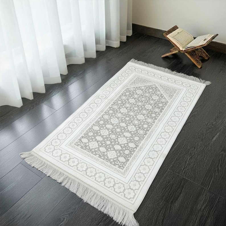 May include: A rectangular prayer rug with a floral pattern in shades of gray and white. The rug has a decorative border and fringe on the ends. A wooden book stand with an open book is in the background.