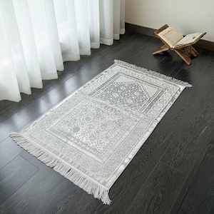 May include: A gray prayer rug with a detailed floral pattern and fringe. The rug is rectangular and placed on a dark wooden floor. A wooden book stand with an open book is in the background.
