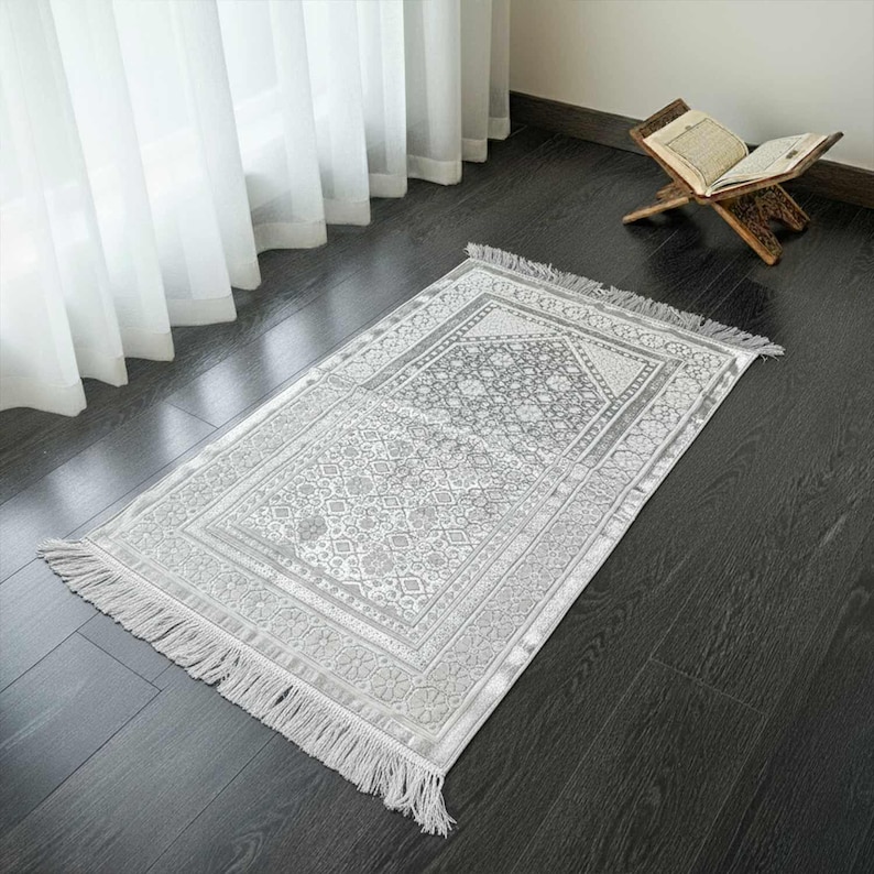 May include: A gray prayer rug with a detailed floral pattern and fringe. The rug is rectangular and placed on a dark wooden floor. A wooden book stand with an open book is in the background.