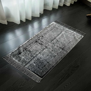 May include: A rectangular gray prayer rug with a detailed floral pattern and fringe trim. The rug is placed on a dark wooden floor, with a window and sheer curtains in the background. The rug's design features a central medallion and border.