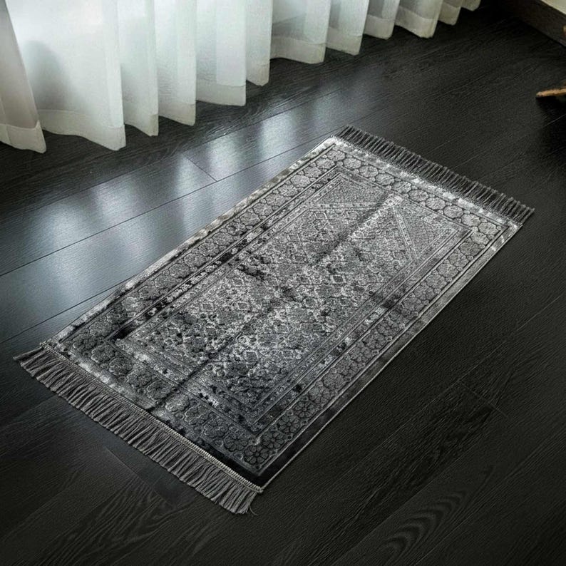 May include: A rectangular gray prayer rug with a detailed floral pattern and fringe trim. The rug is placed on a dark wooden floor, with a window and sheer curtains in the background. The rug's design features a central medallion and border.