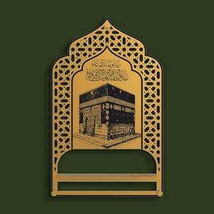 May include: A gold-colored wall-mounted shelf featuring an arched frame with intricate geometric lattice patterns. The central panel displays a black illustration of the Kaaba with Arabic calligraphy above it, set against a dark green background.