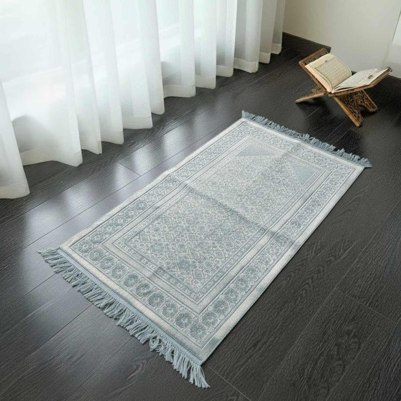 May include: A light blue prayer rug with a detailed floral pattern and fringe trim. The rug is rectangular and lies on a dark wooden floor. A wooden book stand with an open book is visible in the background.