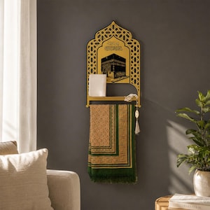 May include: A gold-colored wall-mounted shelf featuring an arched top with intricate geometric cutouts and a black graphic of the Kaaba. A small white book rests on the shelf, and a green and tan patterned prayer rug hangs from the bar below.