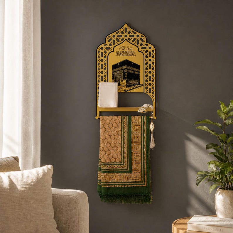 May include: A gold-colored wall-mounted shelf featuring an arched top with intricate geometric cutouts and a black graphic of the Kaaba. A small white book rests on the shelf, and a green and tan patterned prayer rug hangs from the bar below.