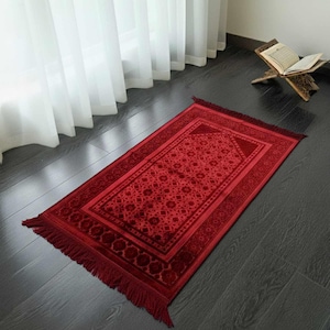 May include: A red prayer rug with a geometric pattern and fringed edges. The rug is rectangular and features a central design with a border. The rug is placed on a dark wooden floor near a window with white curtains.