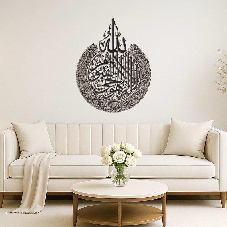 May include: A metallic wall art piece featuring Arabic calligraphy in a circular design. The artwork is in shades of gray and black, with intricate details and a textured appearance. It is mounted on a white wall above a white sofa.