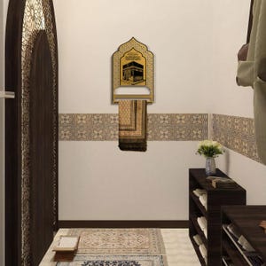 May include: A gold-colored wall-mounted prayer mat hanger featuring an arched top and a central graphic illustration of the Kaaba. A folded brown and beige patterned prayer mat hangs from the horizontal bar of the metal display rack.