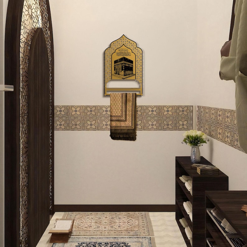 May include: A gold-colored wall-mounted prayer mat hanger featuring an arched top and a central graphic illustration of the Kaaba. A folded brown and beige patterned prayer mat hangs from the horizontal bar of the metal display rack.