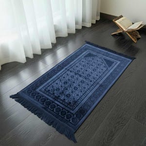 May include: A navy blue prayer rug with a geometric pattern and fringed edges. The rug is rectangular and features a central design surrounded by a border. The rug is placed on a dark wooden floor near a window with white curtains.