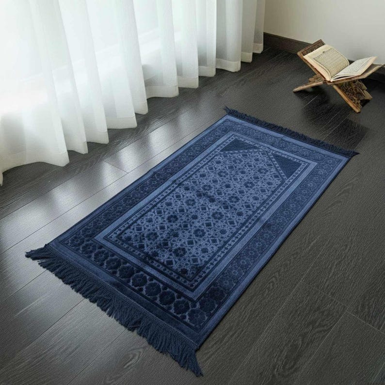 May include: A navy blue prayer rug with a geometric pattern and fringed edges. The rug is rectangular and features a central design surrounded by a border. The rug is placed on a dark wooden floor near a window with white curtains.