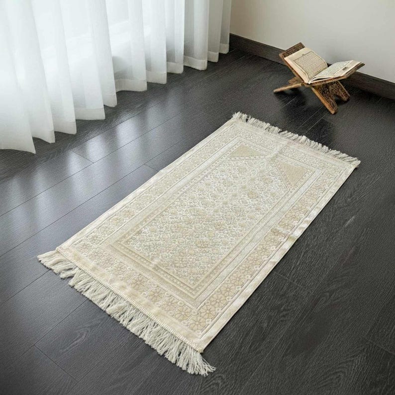 May include: A beige prayer rug with a floral pattern and fringe trim. The rug is rectangular and lies on a dark wood floor. A wooden book stand with an open book is in the background.