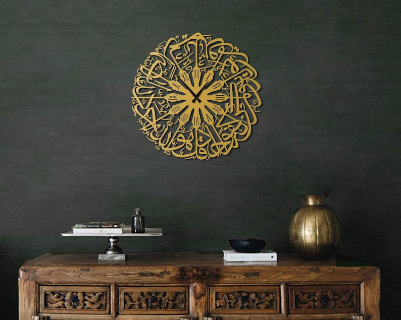 May include: A gold-colored, circular wall clock featuring intricate Arabic calligraphy. The clock hands are black, and the design is mounted on a dark green wall. The clock is a decorative piece, adding a touch of elegance to any room.