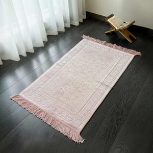 May include: A rectangular prayer rug in a soft pink hue, featuring an intricate floral pattern within a bordered design. The rug has fringed edges and is placed on a dark wooden floor. A wooden book stand with an open book is visible in the background.