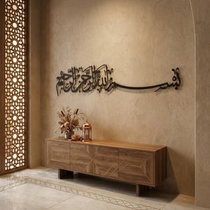 Op de afbeelding: Een zwart metalen wanddecoratie met ingewikkelde Arabische kalligrafie gemonteerd op een getextureerde beige muur boven een lang houten dressoir met vier deuren en geometrische goudkleurige lijnpatronen op de voorpanelen.