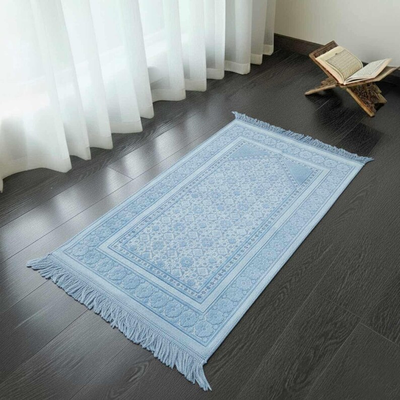 May include: A light blue prayer rug with a floral pattern and fringe. The rug is rectangular and has a detailed border. It is placed on a dark wooden floor near a window with white curtains. A wooden book stand with an open book is visible.