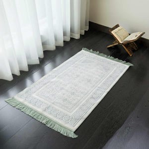 May include: A rectangular prayer rug with a light gray and white floral pattern and a green fringe. The rug is placed on a dark wooden floor near a window with white curtains and a wooden book stand.