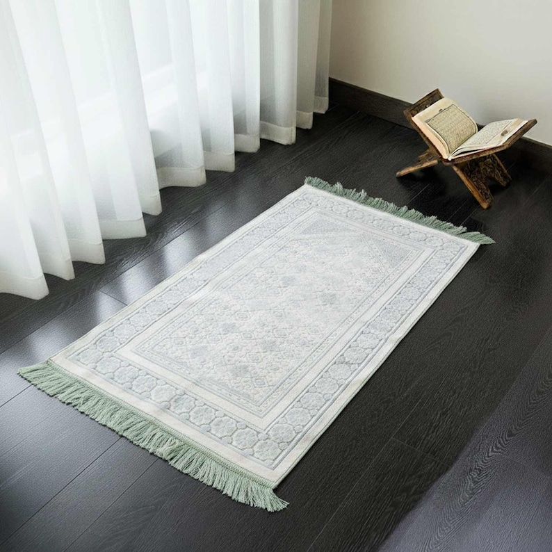 May include: A rectangular prayer rug with a light gray and white floral pattern and a green fringe. The rug is placed on a dark wooden floor near a window with white curtains and a wooden book stand.