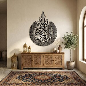 May include: A large, black metal wall art piece featuring Arabic calligraphy, mounted on a beige wall. The artwork is circular with intricate details and is displayed above a wooden cabinet. The room includes a patterned rug, lanterns, and a potted plant.