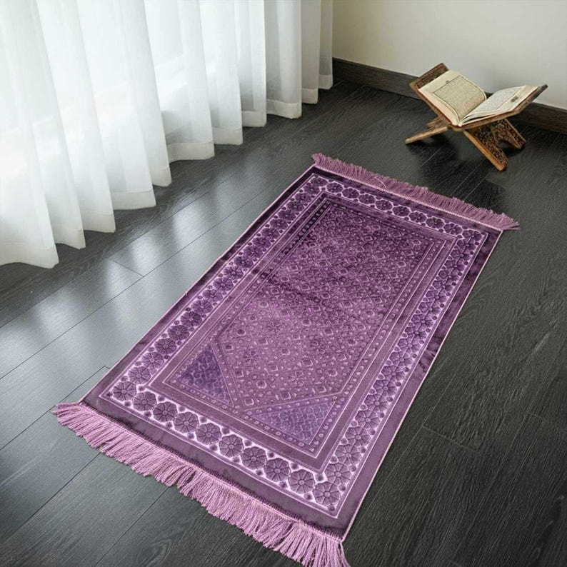 May include: A rectangular prayer rug in shades of purple with a floral border and fringe. The rug is placed on a dark wooden floor, with a wooden book stand holding an open book in the background.