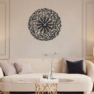 May include: A black, circular wall clock featuring Arabic calligraphy. The clock's hands are black and point to the current time. The clock is mounted on a beige wall above a cream-colored sofa with pillows. The room is decorated with lamps and a coffee table.
