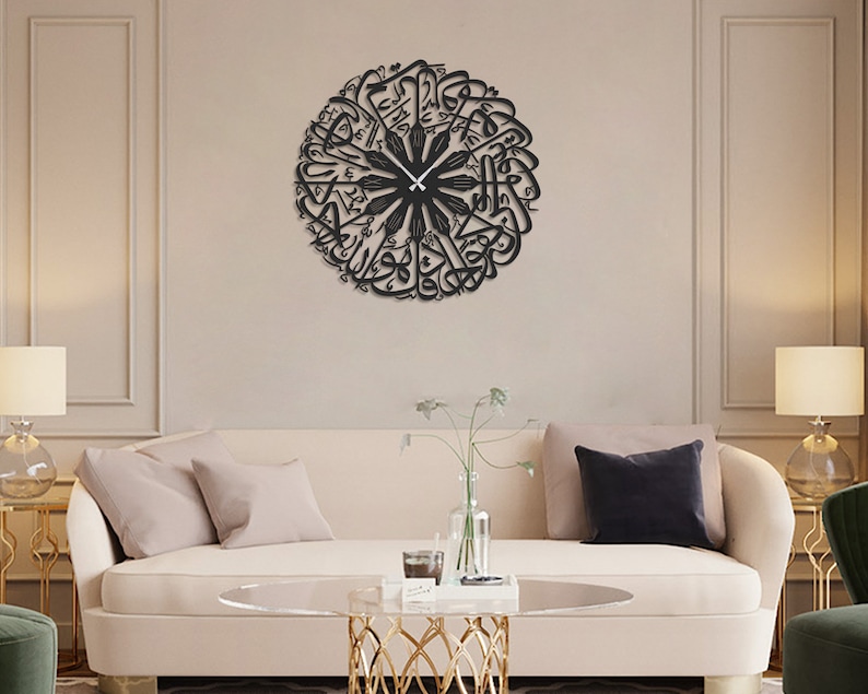 May include: A black, circular wall clock featuring Arabic calligraphy. The clock's hands are black and point to the current time. The clock is mounted on a beige wall above a cream-colored sofa with pillows. The room is decorated with lamps and a coffee table.