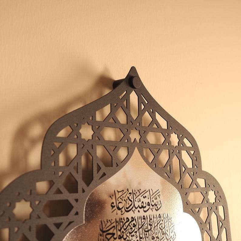 May include: A matte black metal wall hanging featuring an intricate geometric lattice pattern surrounding a central silver-toned panel. The silver panel displays black Arabic calligraphy script. The piece has a pointed arch shape at the top.