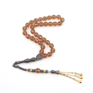 May include: A string of prayer beads featuring translucent, faceted amber-colored glass beads threaded on a green cord. The strand includes a decorative silver-toned metal shield-shaped divider, a textured metal handle, and three gold-toned dangling chains.