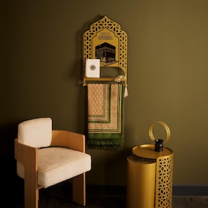 May include: A gold-colored wall-mounted display shelf featuring an arched top with intricate geometric cutouts, a depiction of the Kaaba, and Arabic calligraphy. A green and beige prayer rug hangs from the shelf, with a small white book placed on it.