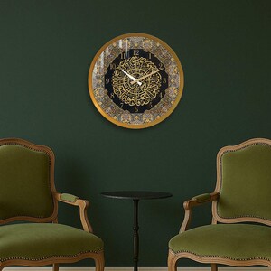 May include: A gold framed clock with Arabic numerals and intricate calligraphy on a black background. The clock is hanging on a dark green wall between two green upholstered chairs.
