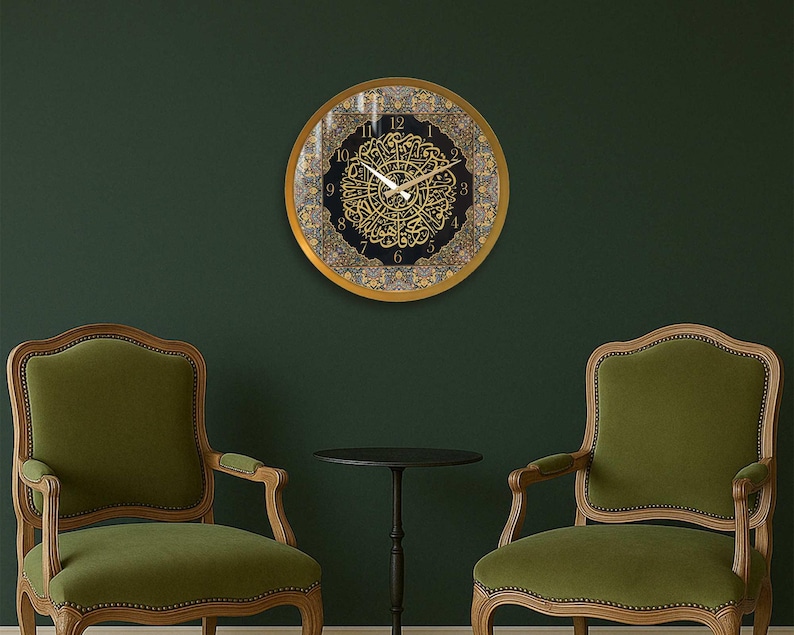May include: A gold framed clock with Arabic numerals and intricate calligraphy on a black background. The clock is hanging on a dark green wall between two green upholstered chairs.