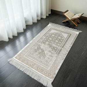 May include: A rectangular prayer rug with a floral pattern in shades of gray and beige. The rug has a decorative border and fringe on the short ends. The rug is placed on a dark wooden floor.