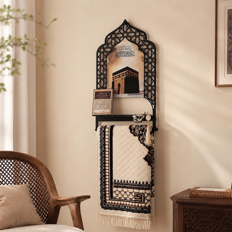 May include: A black metal wall-mounted rack with an arched top featuring geometric lattice patterns. It holds a framed image of the Kaaba, a small book titled Islami Tasarim, prayer beads, and a cream-colored prayer rug with black borders.