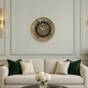 May include: A gold framed clock with black and gold Arabic numerals and a decorative design. The clock is hanging on a white wall with gold trim.