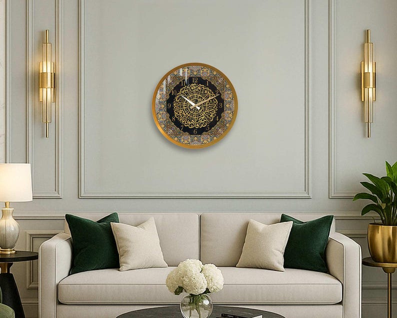 May include: A gold framed clock with black and gold Arabic numerals and a decorative design. The clock is hanging on a white wall with gold trim.
