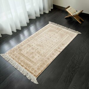 May include: A beige prayer rug with a detailed floral pattern and fringed edges. The rug is rectangular and lies on a dark wooden floor. A wooden book stand with an open book is in the background.