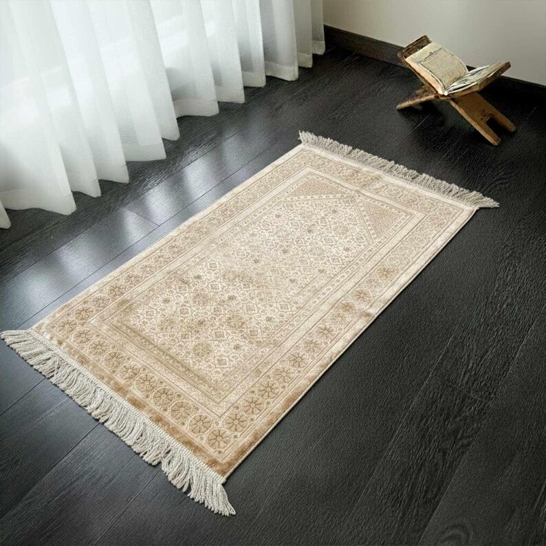 May include: A beige prayer rug with a detailed floral pattern and fringed edges. The rug is rectangular and lies on a dark wooden floor. A wooden book stand with an open book is in the background.