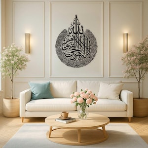 May include: A black metal Islamic calligraphy art piece hangs above a white sofa. The artwork is circular with intricate script. The living room features a round wooden coffee table, a vase of pink flowers, and two potted trees.