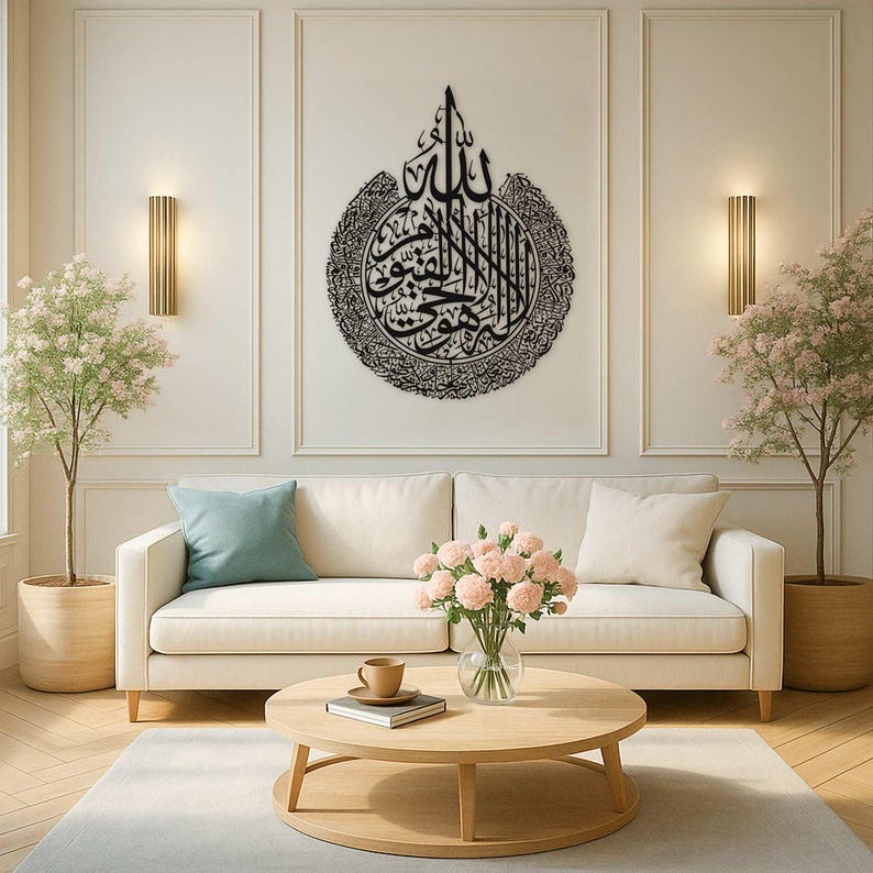 May include: A black metal Islamic calligraphy art piece hangs above a white sofa. The artwork is circular with intricate script. The living room features a round wooden coffee table, a vase of pink flowers, and two potted trees.
