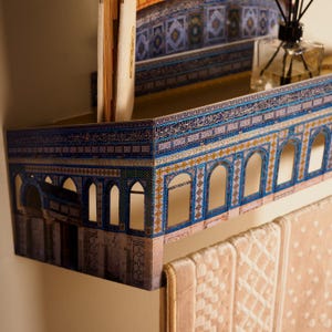 Pu&ograve; includere: Una mensola decorativa con un design architettonico dettagliato con accenti blu, oro e bianco. La mensola presenta aperture ad arco e motivi intricati, che ricordano una struttura storica. Un libro e un diffusore di fragranze sono posizionati sulla mensola, con un asciugamano beige appeso sotto.