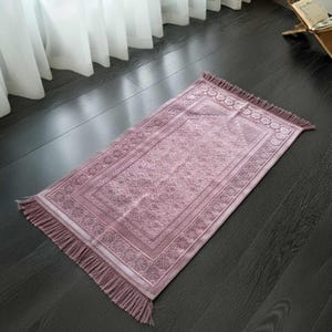May include: A rectangular prayer rug in a soft mauve color with intricate floral and geometric patterns. The rug has a decorative border and fringe on the short ends. The rug is placed on a dark wooden floor.