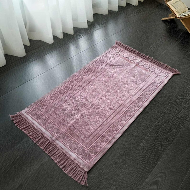 May include: A rectangular prayer rug in a soft mauve color with intricate floral and geometric patterns. The rug has a decorative border and fringe on the short ends. The rug is placed on a dark wooden floor.